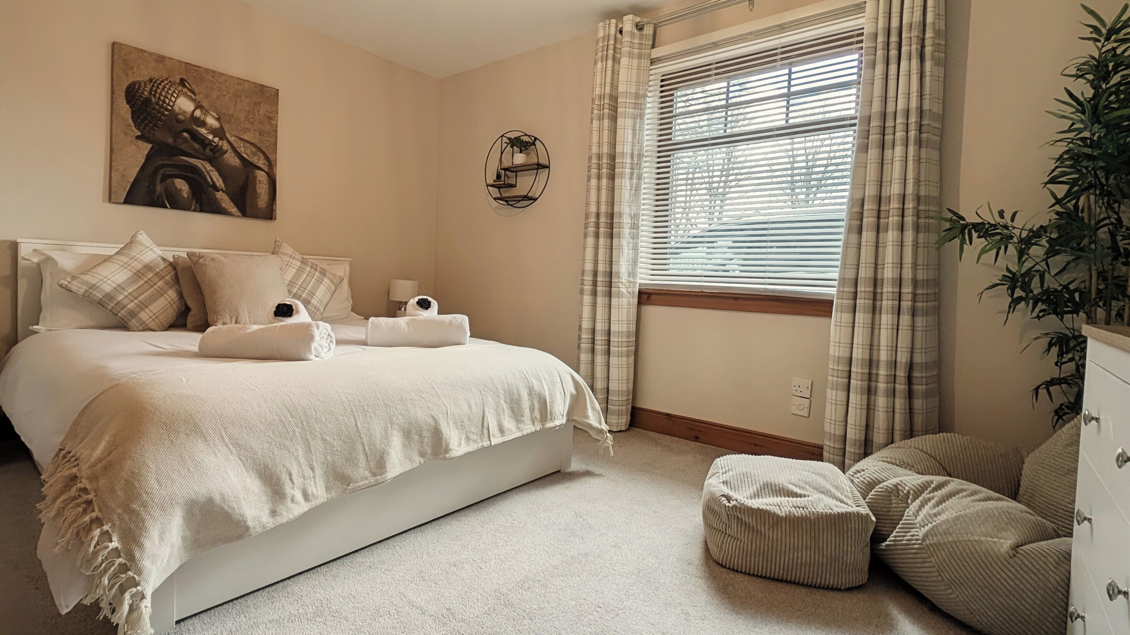 A bedroom with a bed, pillows, a bedside table with a lamp, a window with blinds, a wall-mounted shelf with a plant, and a painting.