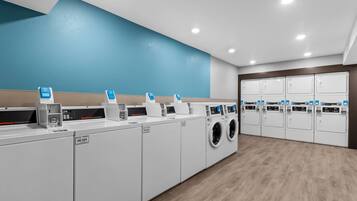 A commercial laundry facility with multiple washing machines and dryers, a blue wall, and wooden flooring.