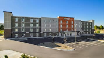 A multi-story hotel with a central entrance, parking lot, and a landscaped area with small trees.
