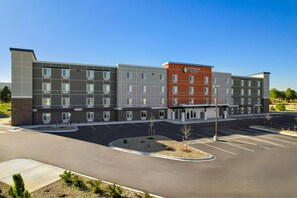 A multi-story hotel with a central entrance, parking lot, and a landscaped area with small trees.