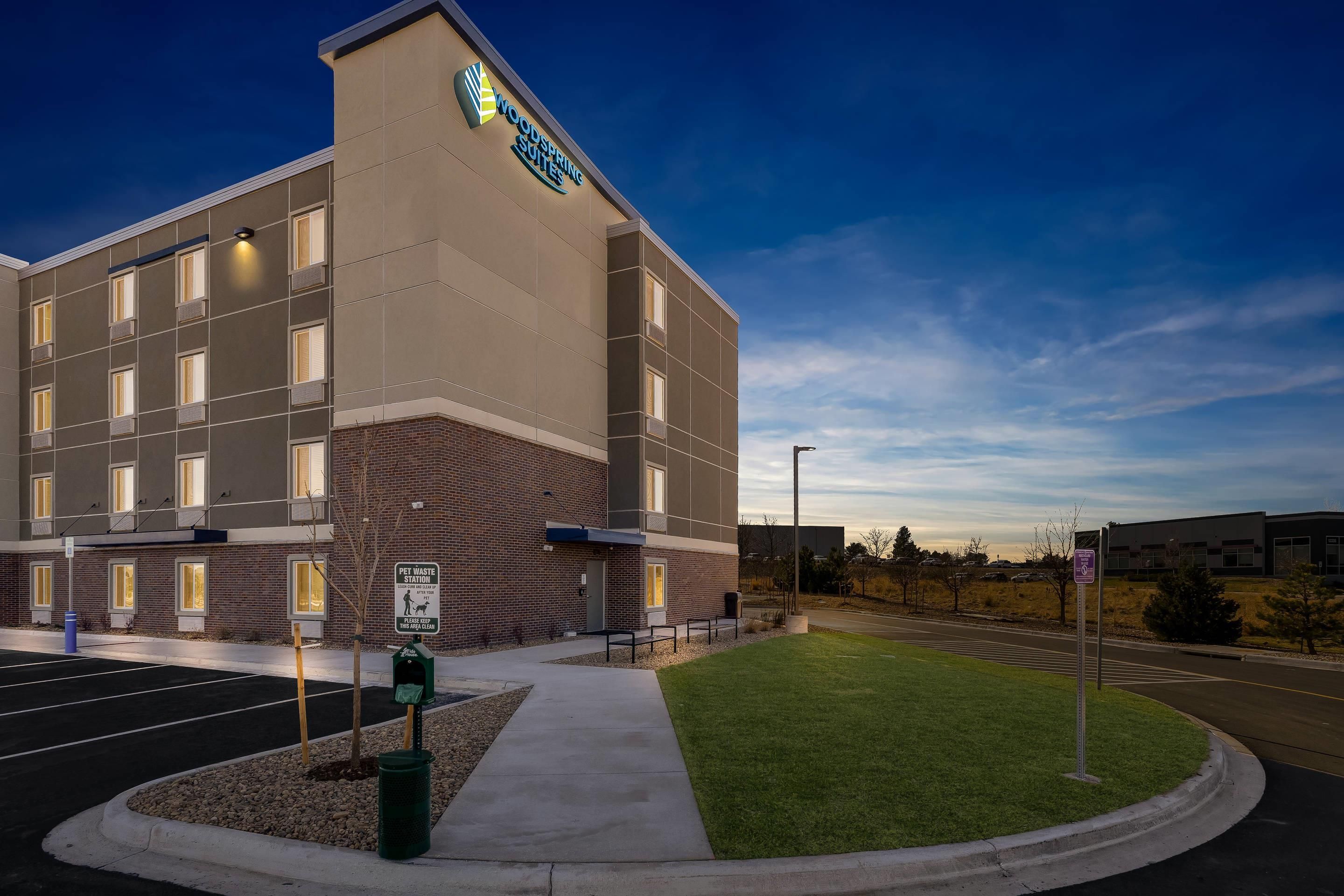 A hotel with a sign for WoodSpring Suites, a pet waste station, and a parking lot.