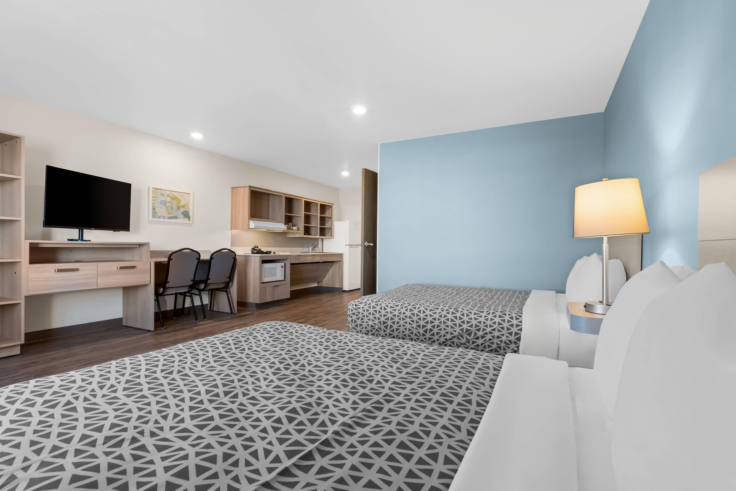 A modern hotel room with a desk, kitchenette, two beds with patterned bedding, and a TV.