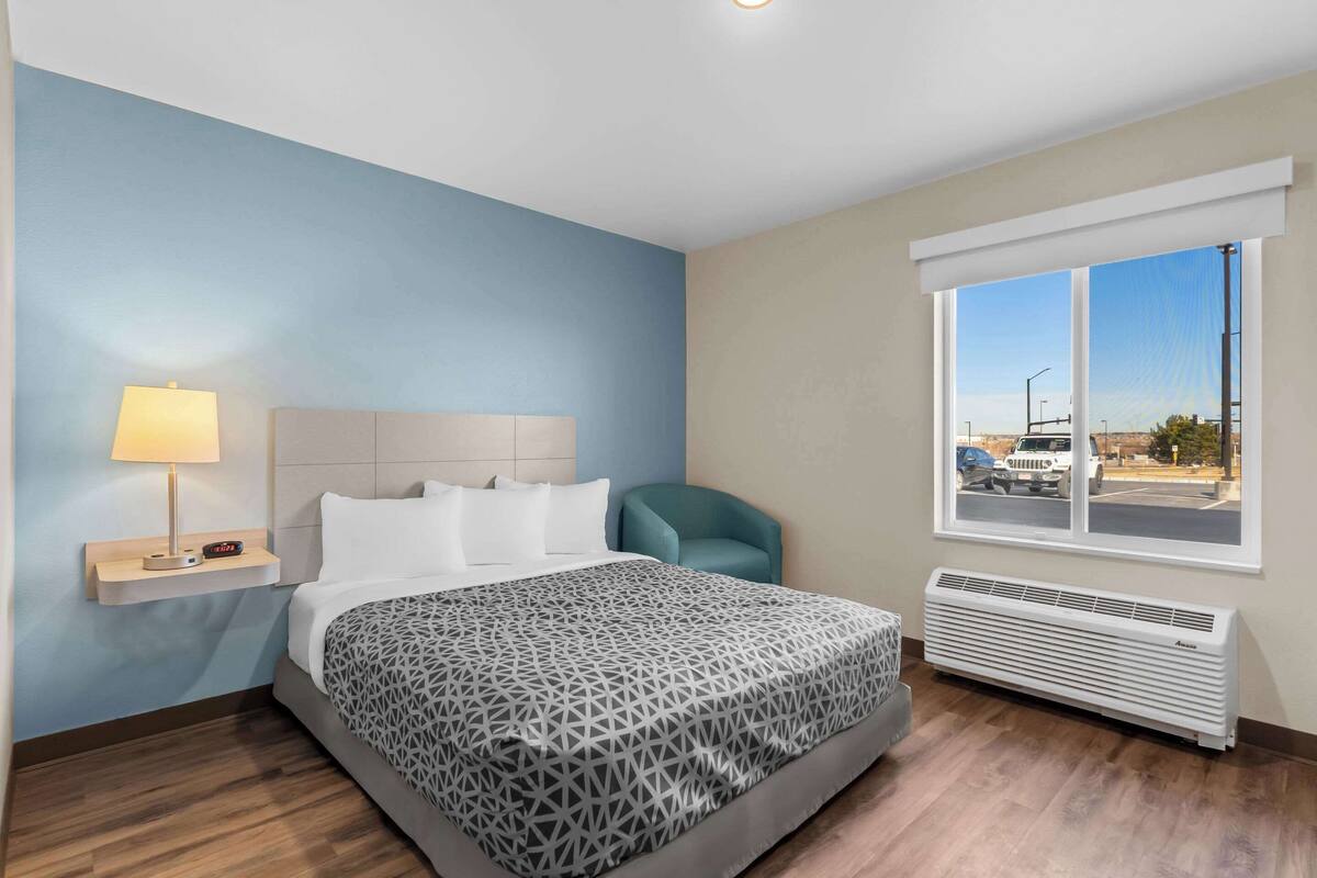 Una habitación de hotel con cama, mesita de noche con lámpara, una silla, ventana con vista a la calle y aire acondicionado instalado en la pared.