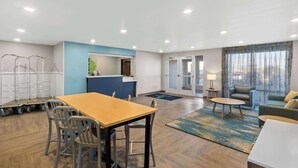 A modern hotel room with a wooden dining table, chairs, a sofa, and a small round table. There is a kitchen area with a counter and cabinets, and a large window with curtains. - WoodSpring Suites Centennial - Denver Tech Center (Centennial)