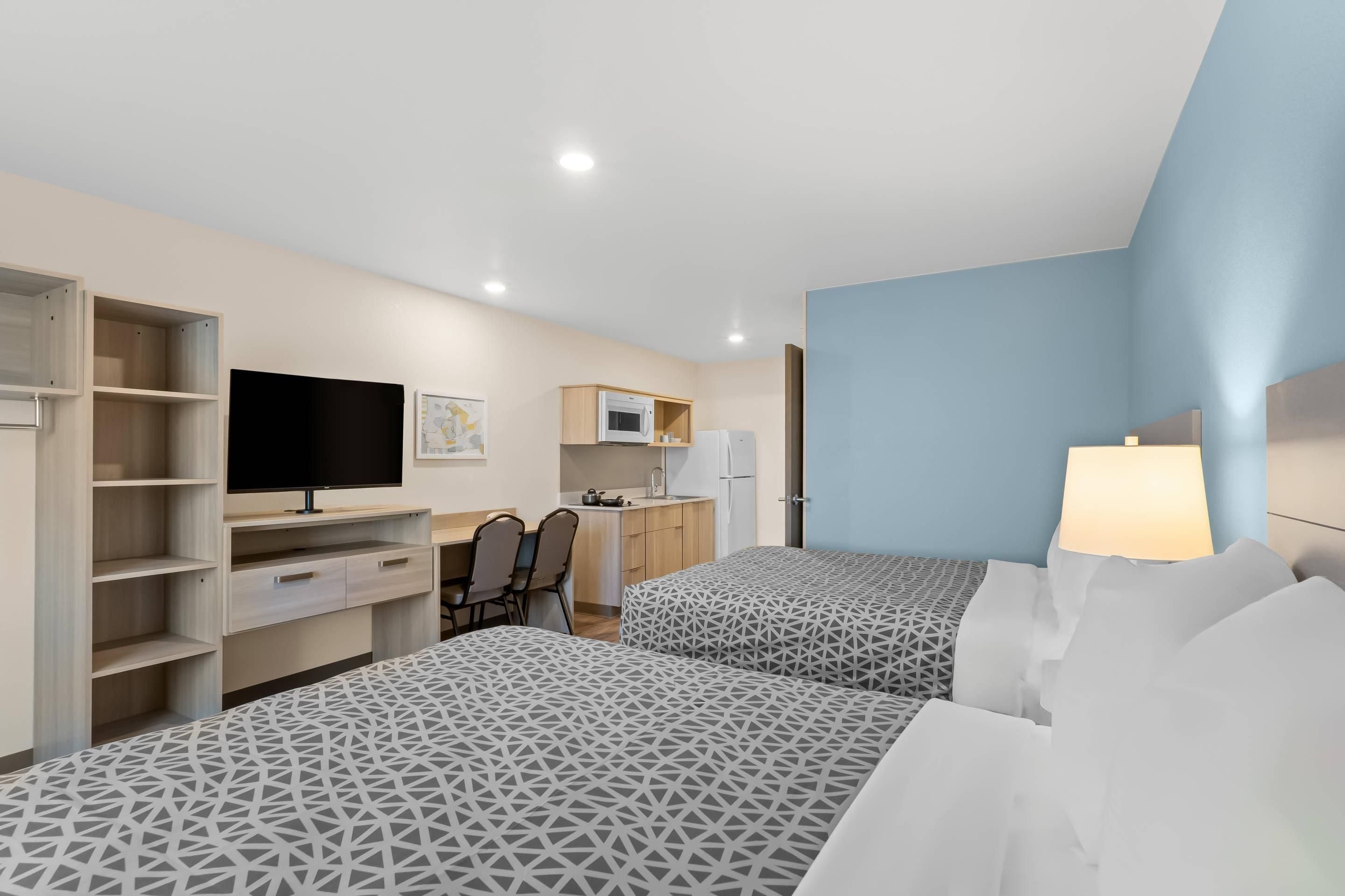 A hotel room with two beds, a desk, a television, and a kitchenette.