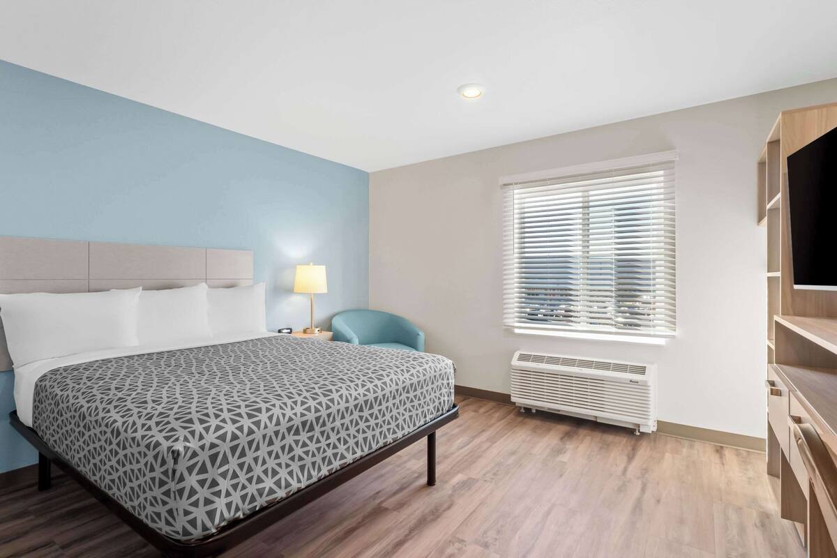 Habitación de hotel con cama, escritorio, silla, televisor y ventana con persianas.