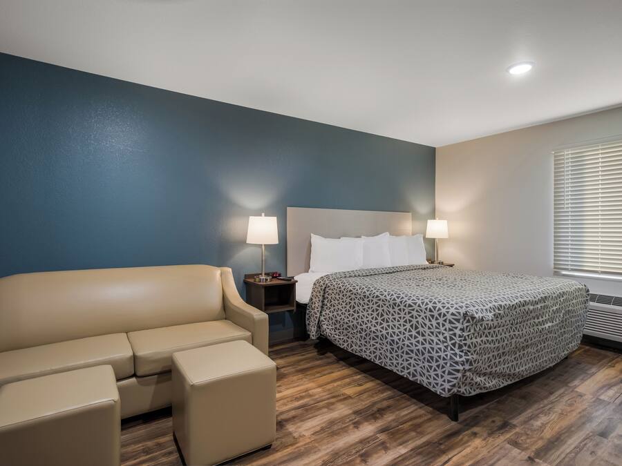 Una habitación de hotel con cama, sofá, dos mesitas de noche con lámparas y una ventana con persianas.