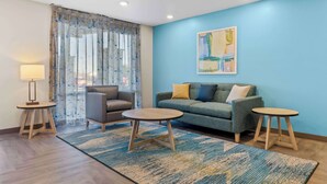 A modern living room with a grey sofa, two round wooden tables, a floor lamp, and a blue accent wall. - WoodSpring Suites Centennial - Denver Tech Center (Centennial)