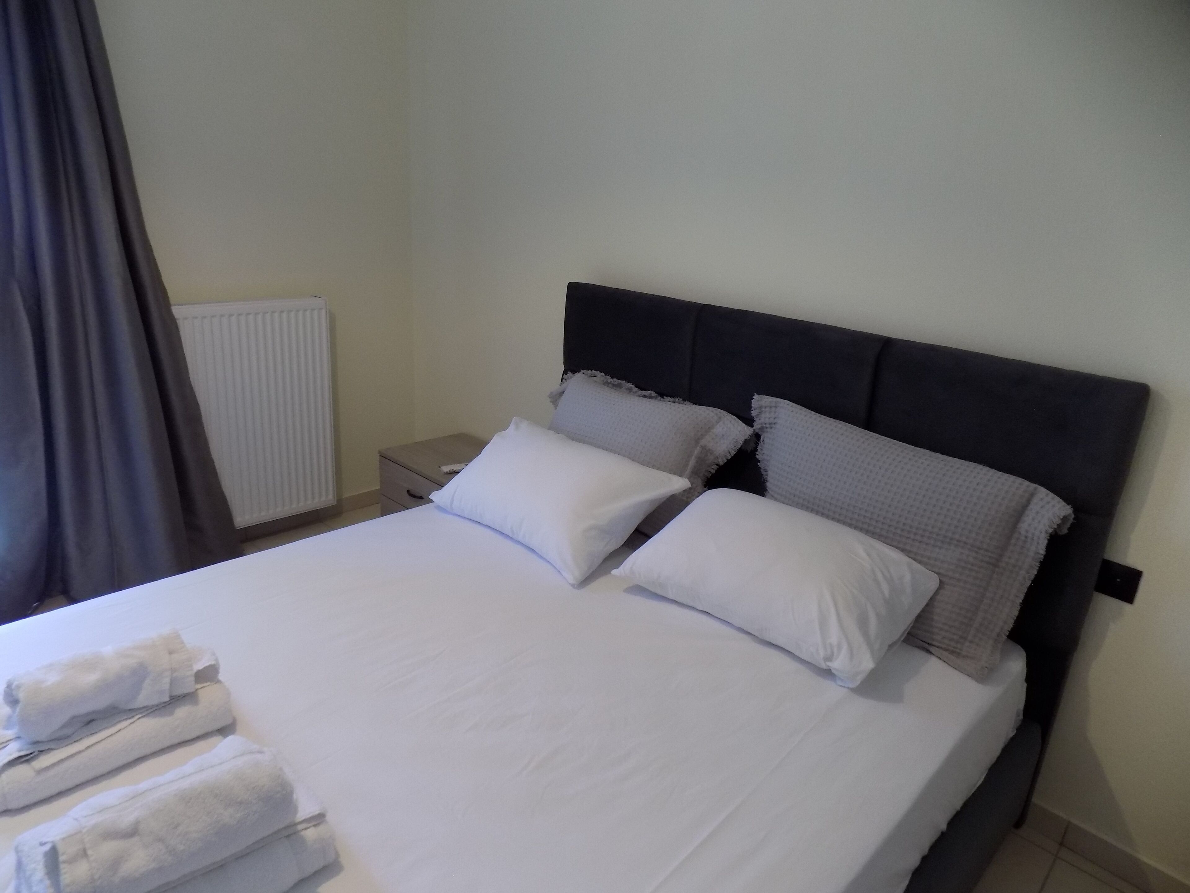 A neatly made bed with white linens and pillows, a bedside table with a lamp, and a radiator on the wall.