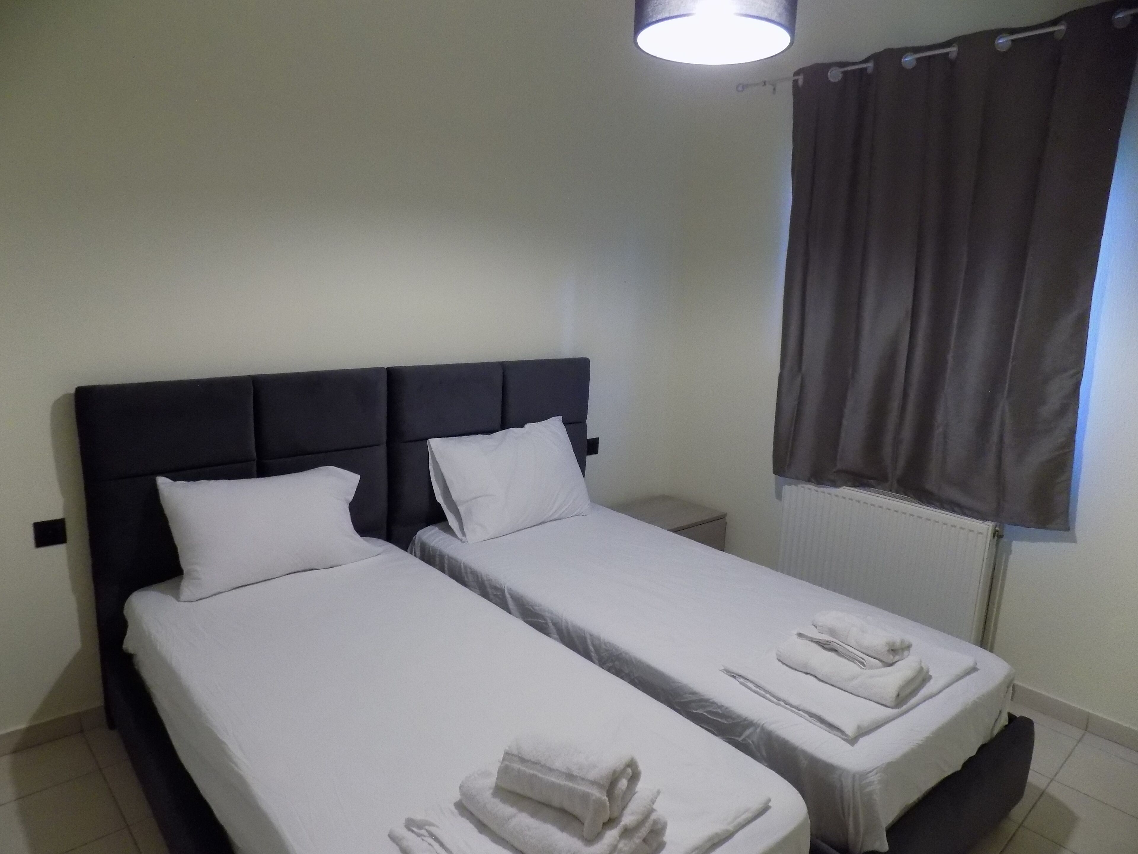 A hotel room with two single beds, each with white linens and pillows, a dark headboard, a small bedside table, and a window with curtains.