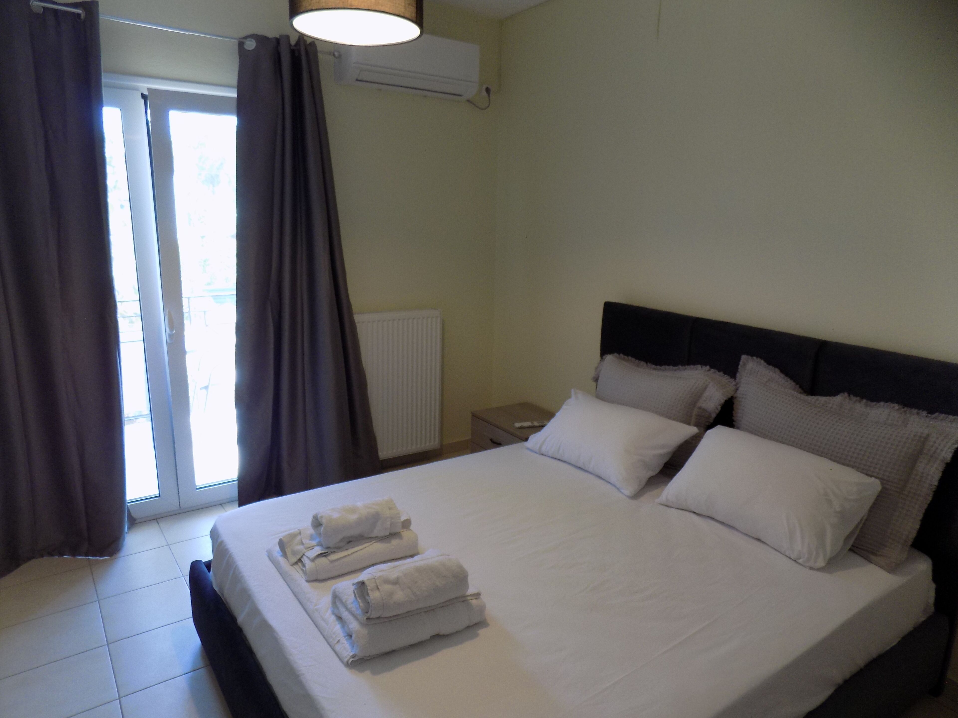 Habitación de hotel con cama, almohadas, mesita de noche, control remoto y ventana con cortinas.