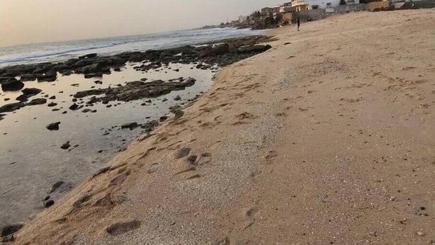 Una playa arenosa con rocas dispersas y huellas, un mar tranquilo y edificios al fondo.