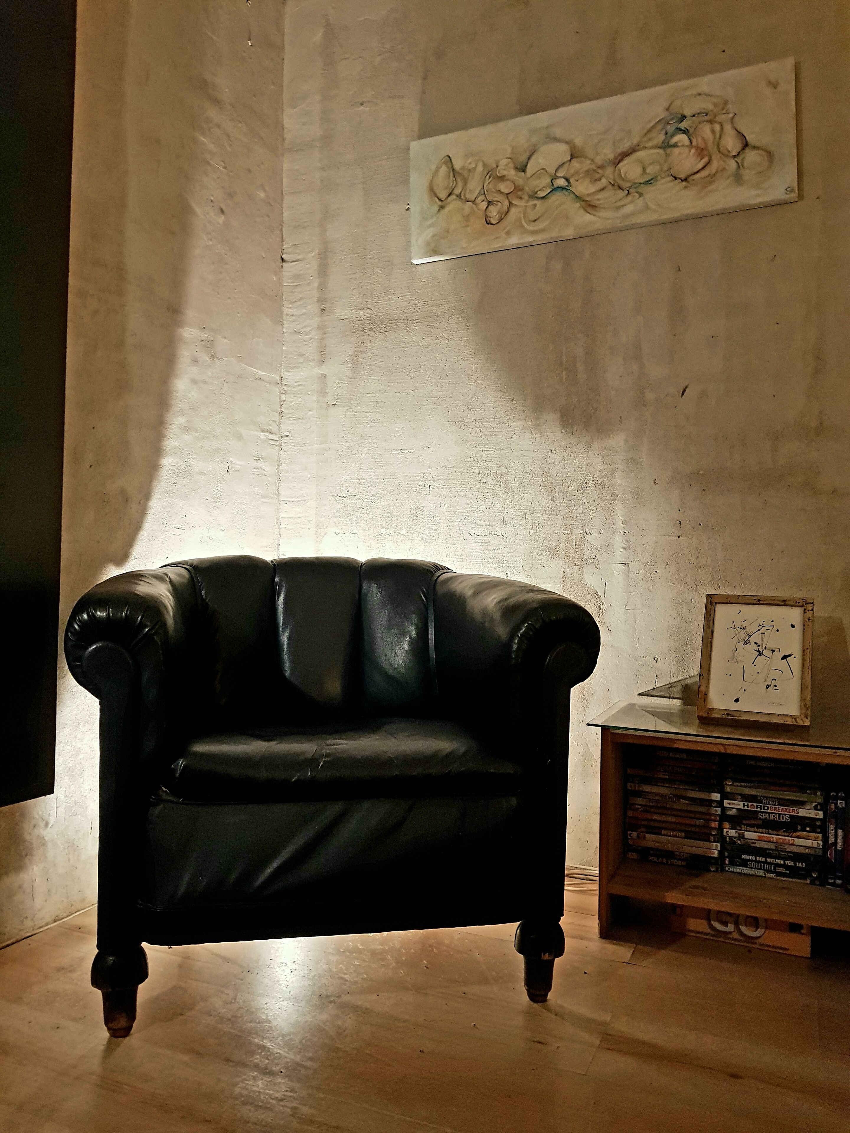 Een zwarte leren bank in een kamer met een betonwand en een houten plank met boeken.