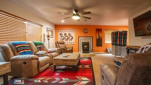 A living room with a sofa, armchair, and coffee table. There is a fireplace, a ceiling fan, and a rug with geometric patterns. - Spacious Route 66 retreat. Golf Course. Pet Friendly. Grand Canyon and Vegas. (Kingman)