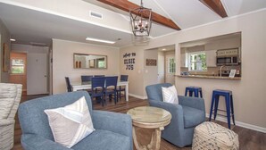 Uma sala de estar espaçosa com sofás azuis, uma mesa de centro redonda e uma área de jantar com mesa branca e cadeiras azuis. É visível uma área de cozinha com uma placa indicando "Lake Martin".