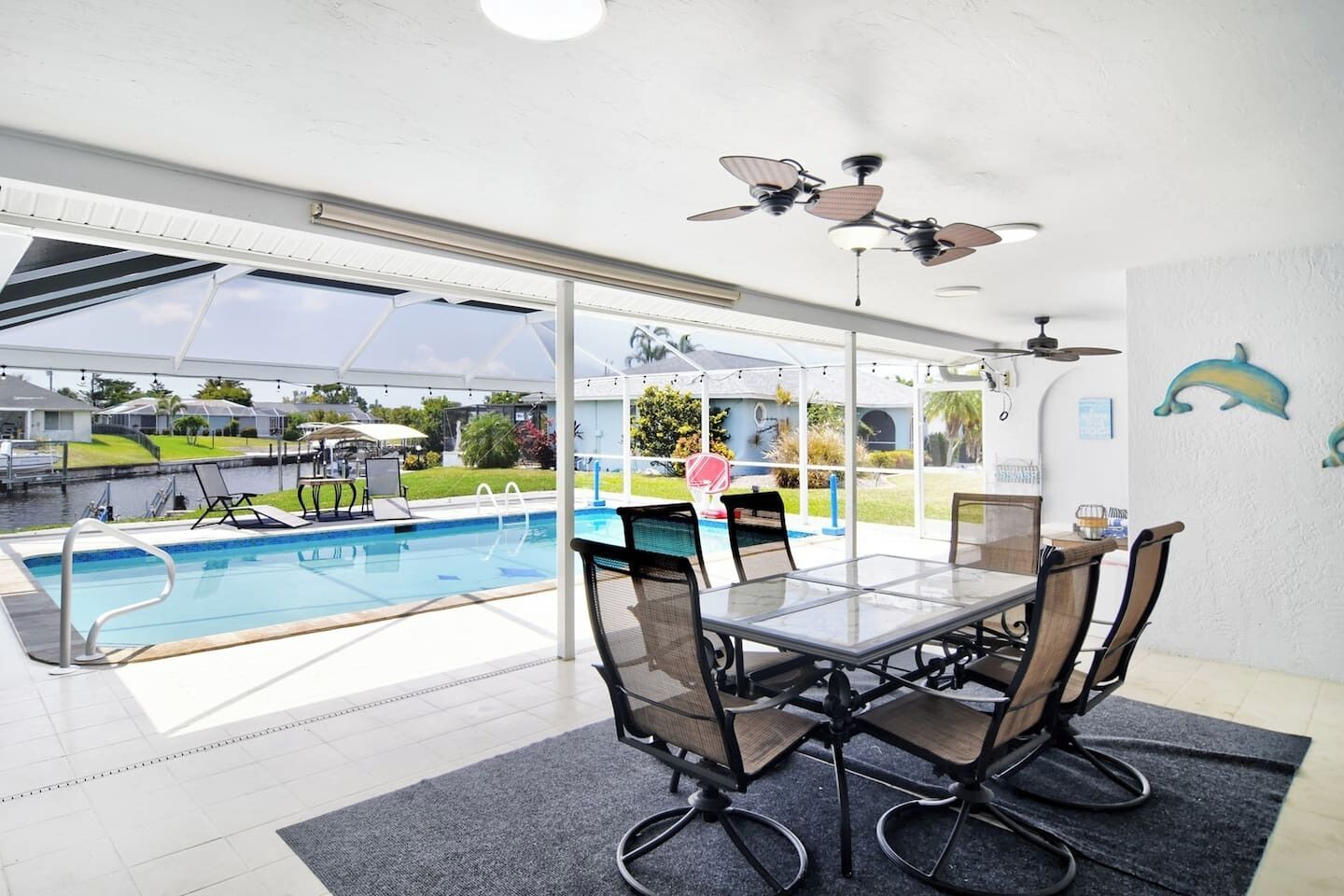 A poolside dining area with a glass table and chairs, a ceiling fan, and a view of a swimming pool and outdoor area.