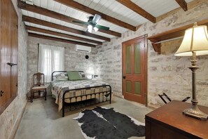 Ein rustikales Schlafzimmer mit sichtbaren Holzbalken, einem Bett mit Quilzudecke, einem Kuhfellteppich, einer Lampe und einer Steinwand.