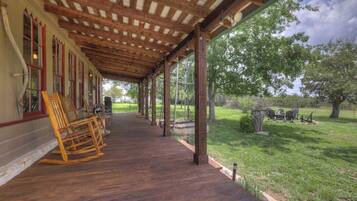 A covered porch with wooden flooring and rocking chairs, overlooking a grassy area with trees and outdoor furniture.