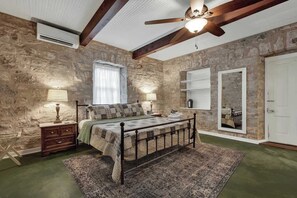 A bedroom with a stone wall, a bed with a metal frame, a wooden nightstand, a floor lamp, a mirror, and a ceiling fan.