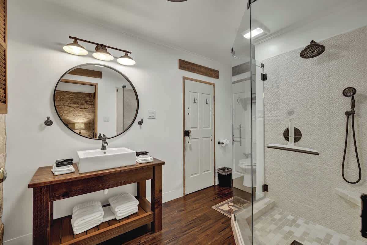 Une salle de bain avec un meuble-lavabo en bois, un miroir rond, un lavabo blanc et une douche séparée par une paroi en verre.