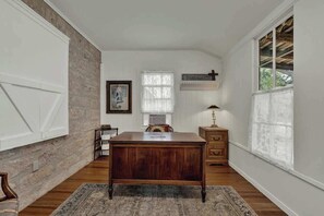 Ein Zimmer mit Schreibtisch aus Holz, einem Stuhl, einem kleinen Tisch, einer Lampe und einem Fenster mit Kruzifix.