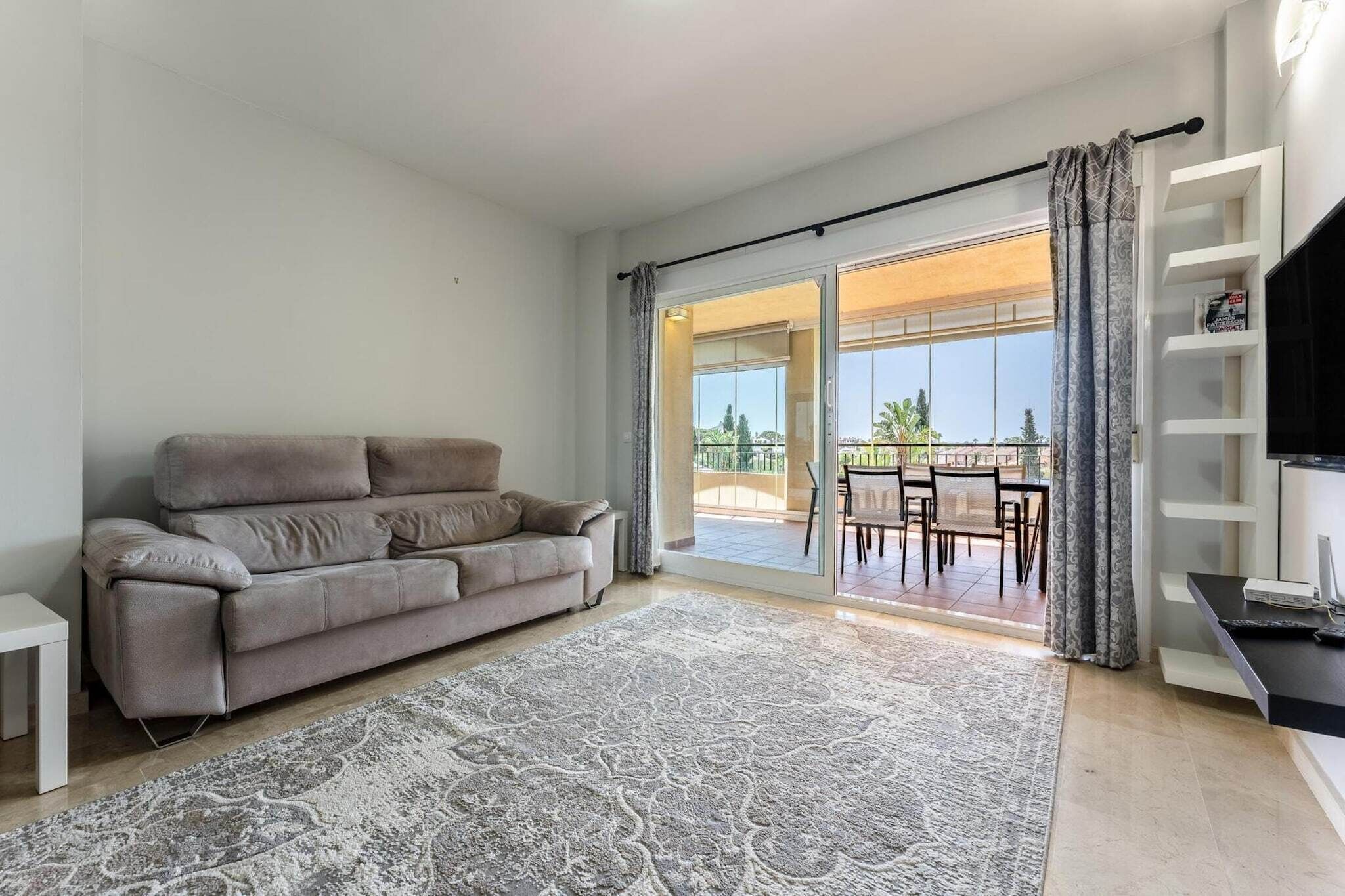 Ein Wohnzimmer mit grauem Sofa, einem gemusterten Teppich, einer TV-Konsole und einer Glastür zum Außenbereich.
