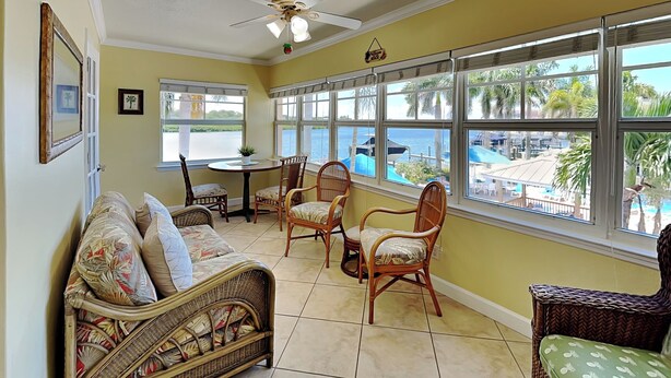 A room with a wicker sofa, wicker chairs, a ceiling fan, and large windows overlooking a pool and palm trees. - Water View Condo Across from Beach + Access to Amenities (Indian Shores)