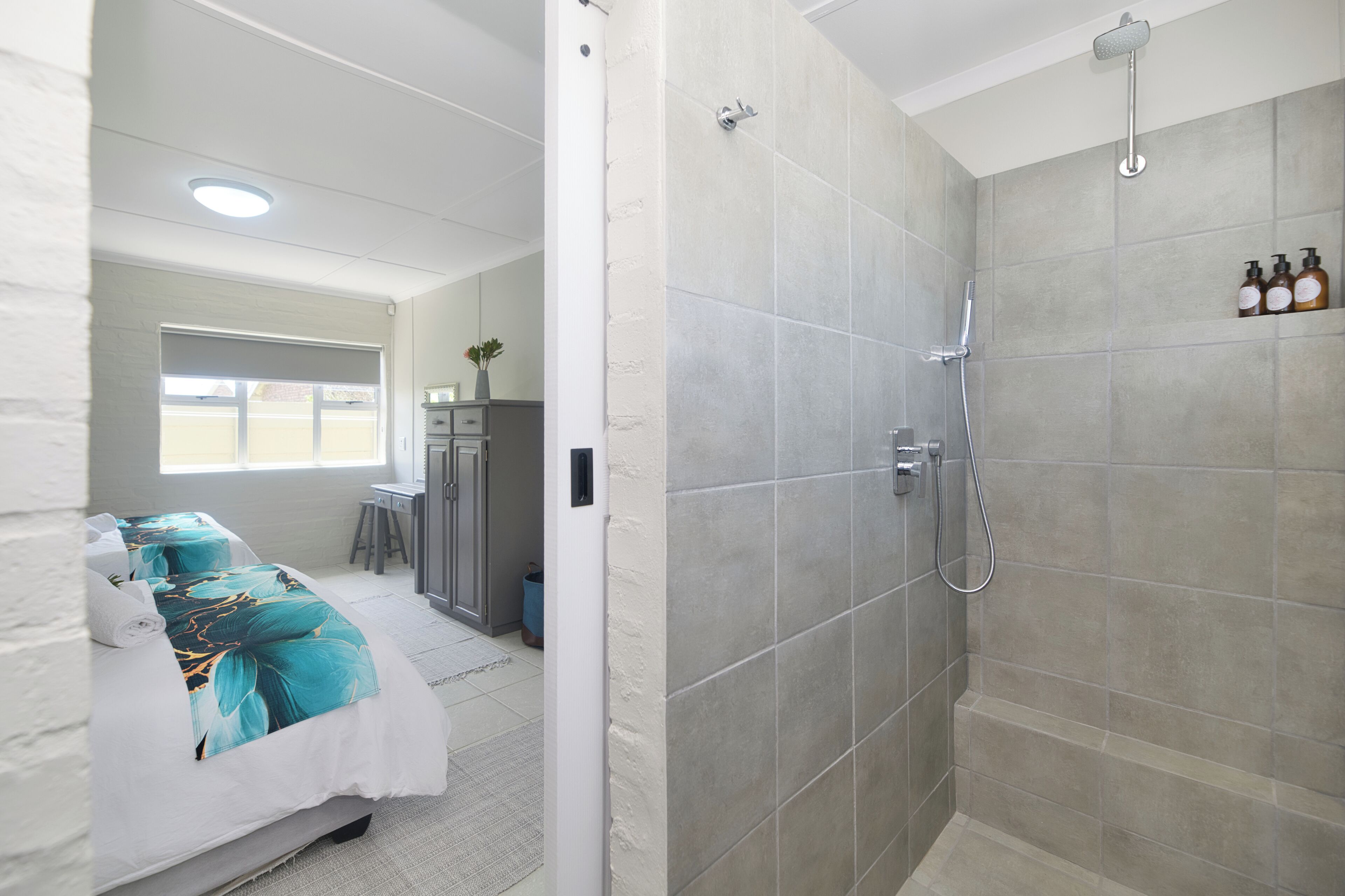 Ein modernes Badezimmer mit Dusche, ein Bett mit floralem Überwurf und ein Fenster, das natürliches Licht hereinlässt.