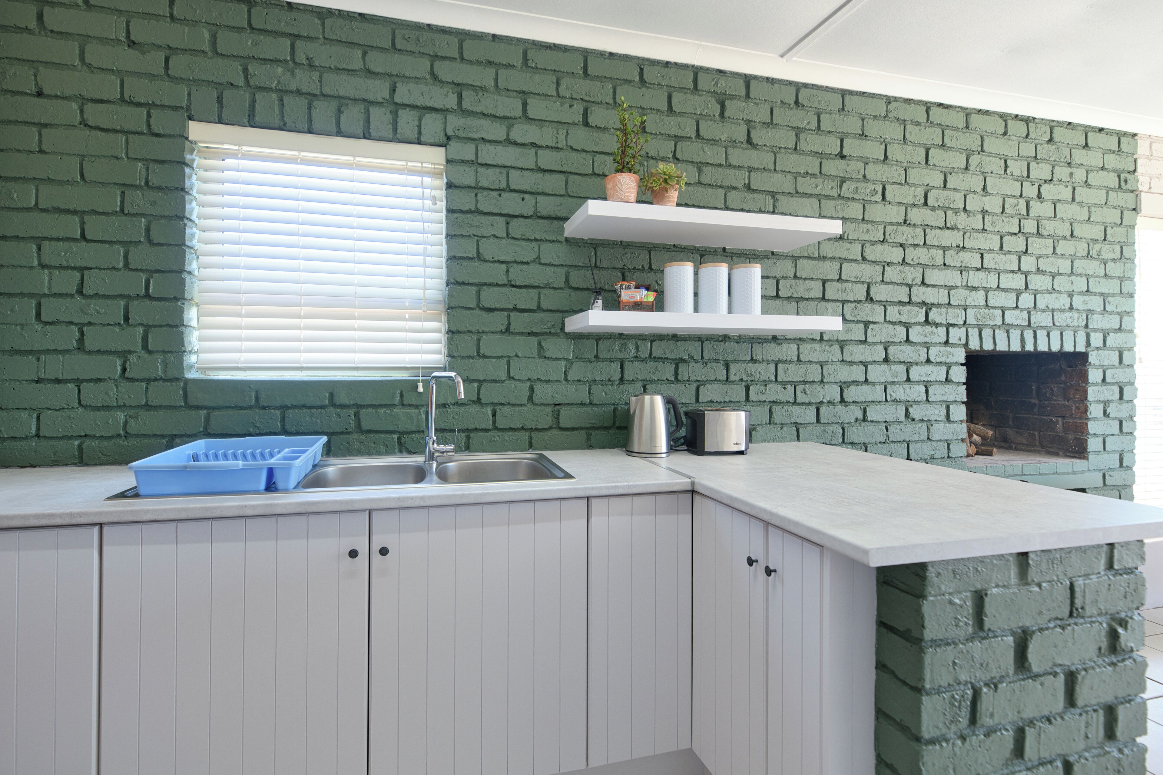 Una cocina con armarios blancos, pared de ladrillo verde, ventana con persianas y chimenea de ladrillo empotrada.