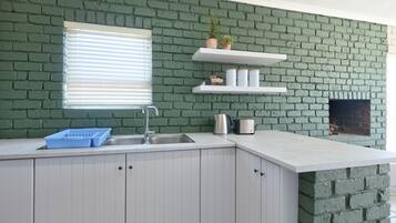 Una cocina con armarios blancos, pared de ladrillo verde, ventana con persianas y chimenea de ladrillo empotrada.