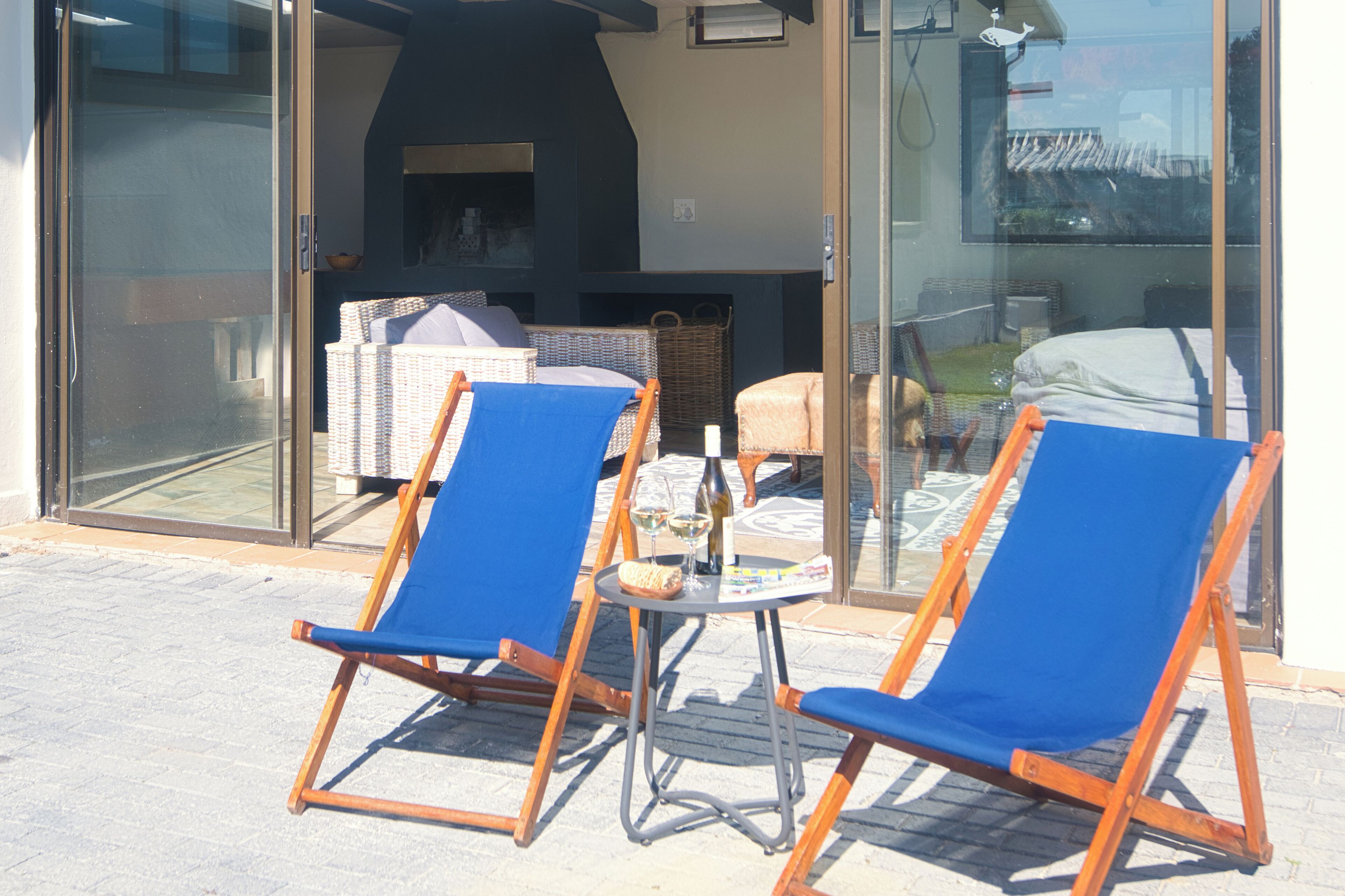 Two blue lounge chairs outside a building with a glass door, a wicker basket, and a small table with a bottle and glass.