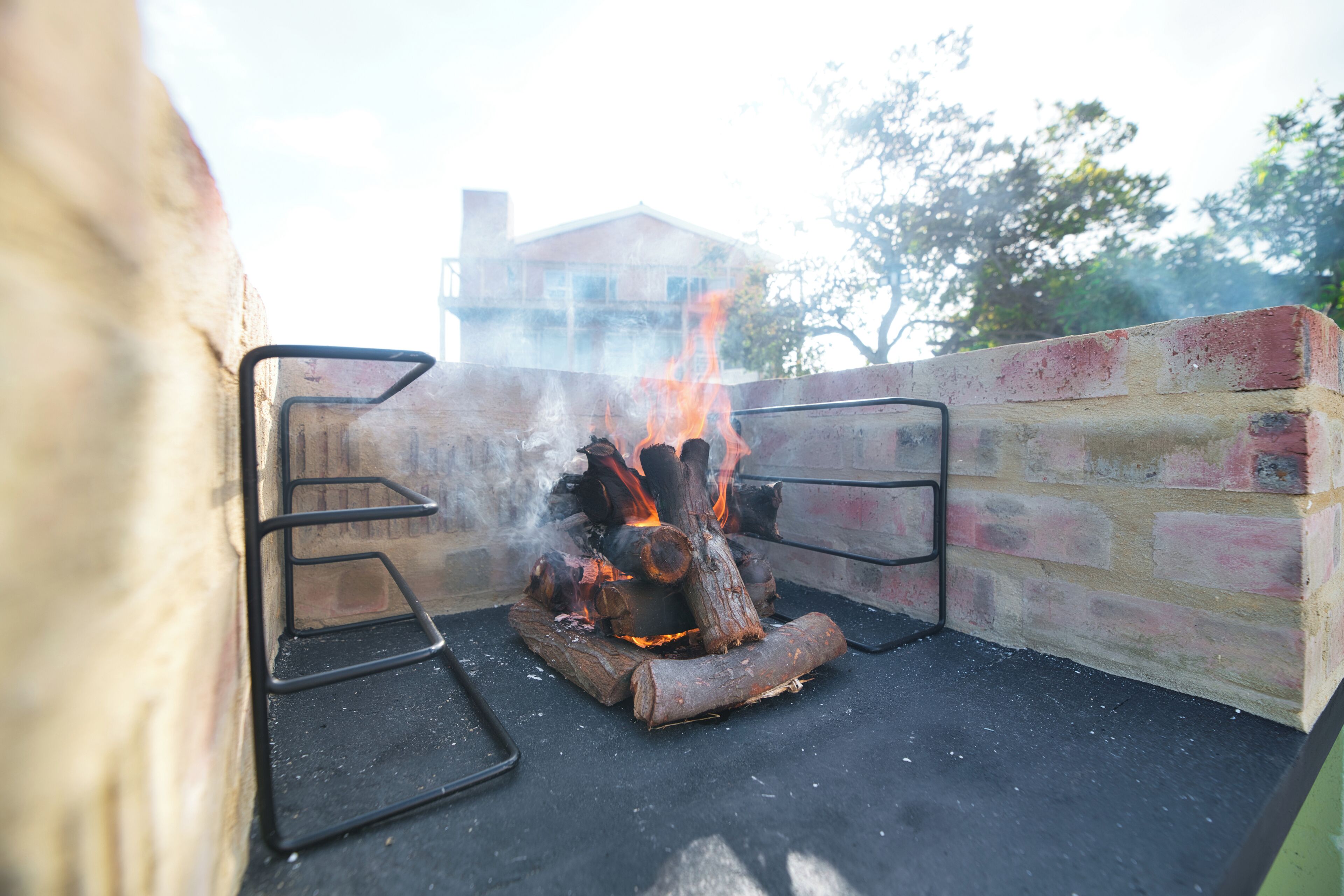 Una parrilla de ladrillo con leña ardiendo y llamas, frente a un muro de ladrillo y una baranda metálica.