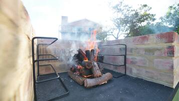 Una parrilla de ladrillo con leña ardiendo y llamas, frente a un muro de ladrillo y una baranda metálica.