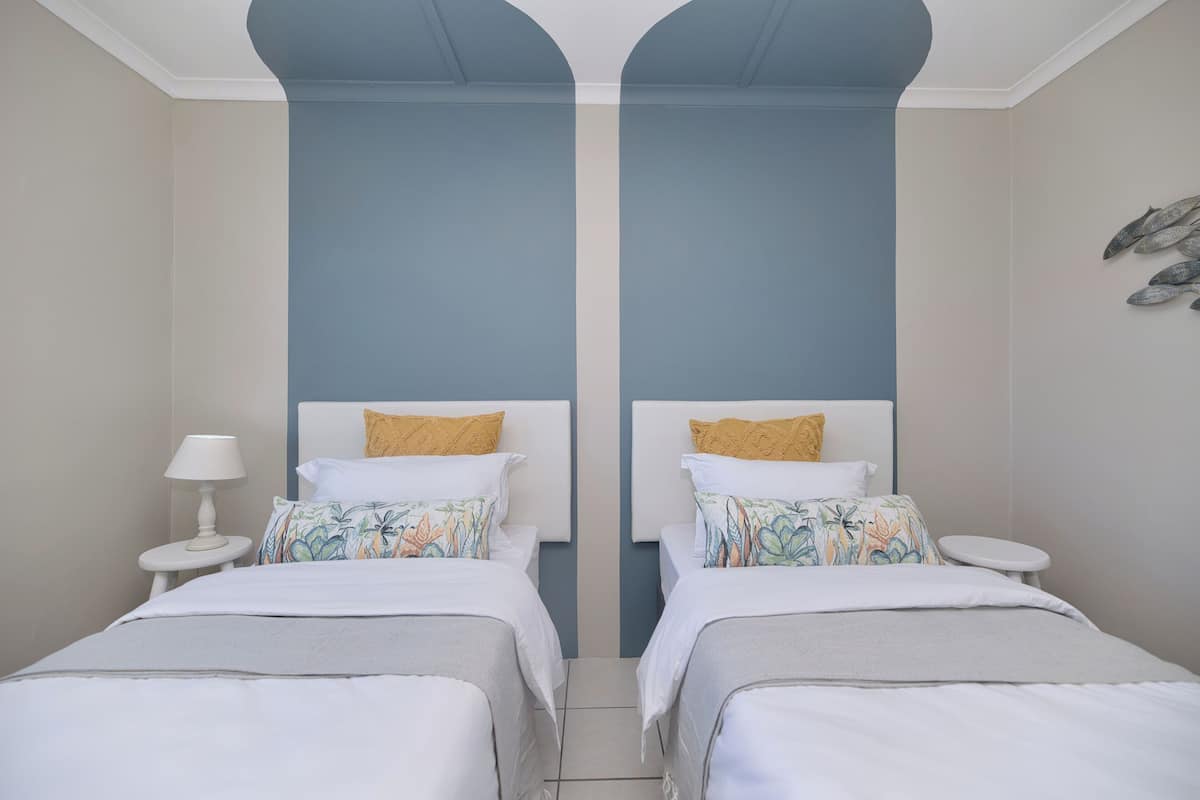 Una habitación con dos camas, cada una con cabecero blanco y almohada estampada. A la izquierda, una mesita pequeña con una lámpara.