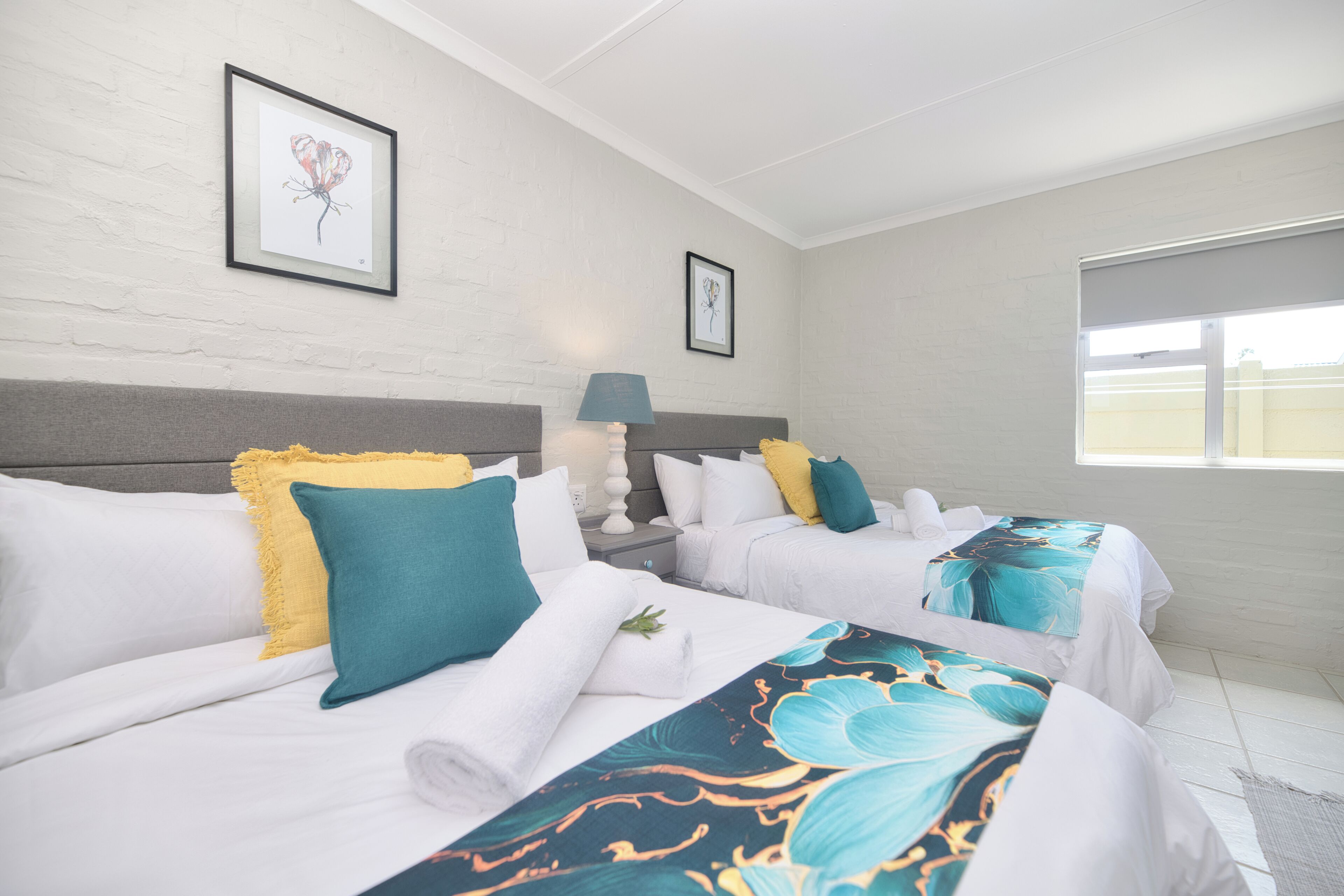 A hotel room with two beds, white bedding, teal and yellow pillows, a nightstand with a lamp, and framed artwork on the walls.