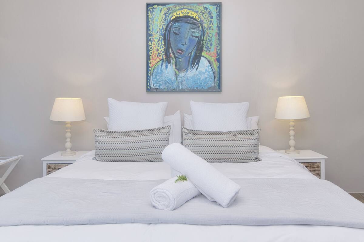 Una cama bien hecha con sábanas blancas, dos mesitas de noche con lámparas y un cuadro sobre la cama.