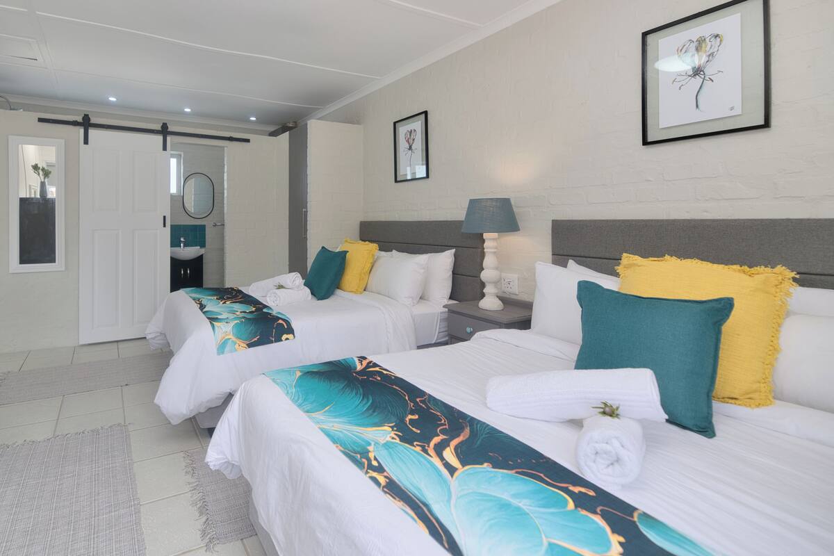 Habitación de hotel con dos camas, una mesita de noche, una lámpara, un cuadro en la pared y un baño visible a través de una puerta abierta.