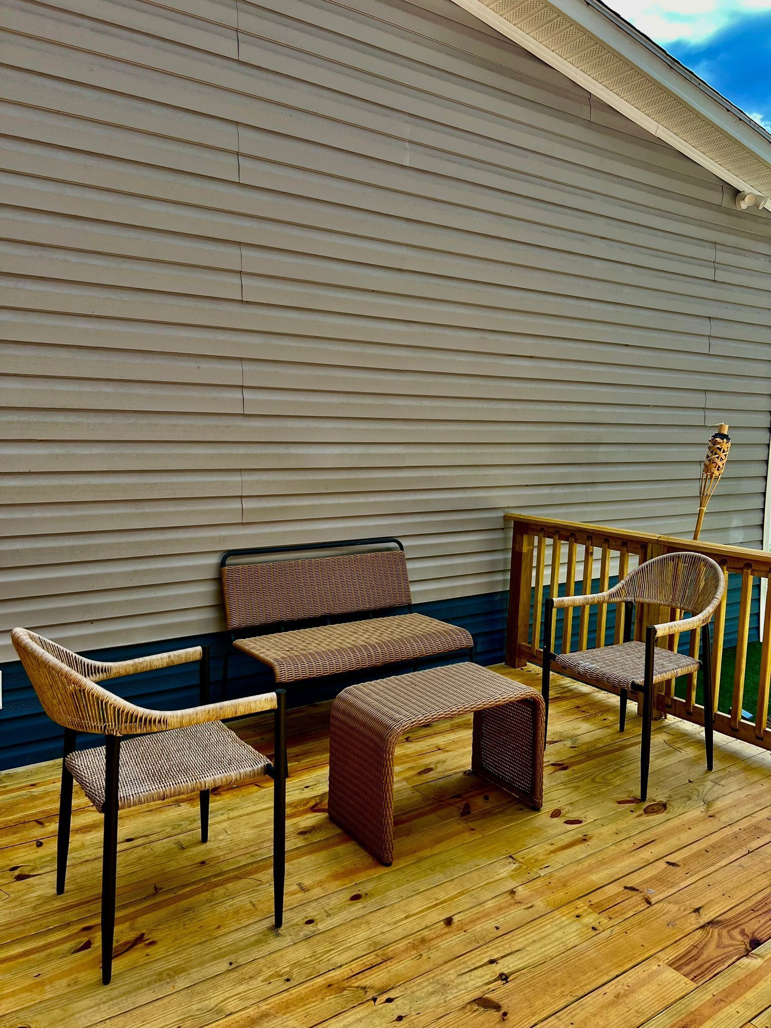 Une terrasse en bois, agrémentée de meubles en osier, dont un banc et des chaises, adossée à un bâtiment dont le revêtement extérieur est constitué de planches disposées horizontalement.