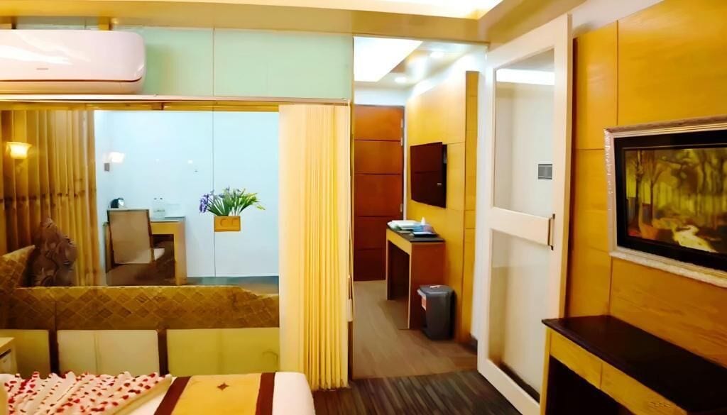 A hotel room with a bed, a desk, a chair, a TV, a plant, and a painting on the wall.