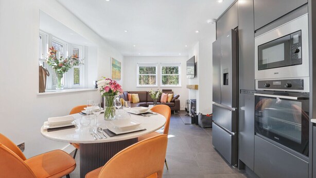 A modern kitchen with stainless steel appliances, a dining area with orange chairs, and a view of the outdoors through large windows.