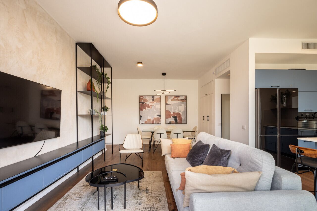 A modern living room with a large flat-screen TV, a grey sofa, a coffee table, and a dining area with white chairs and a table.