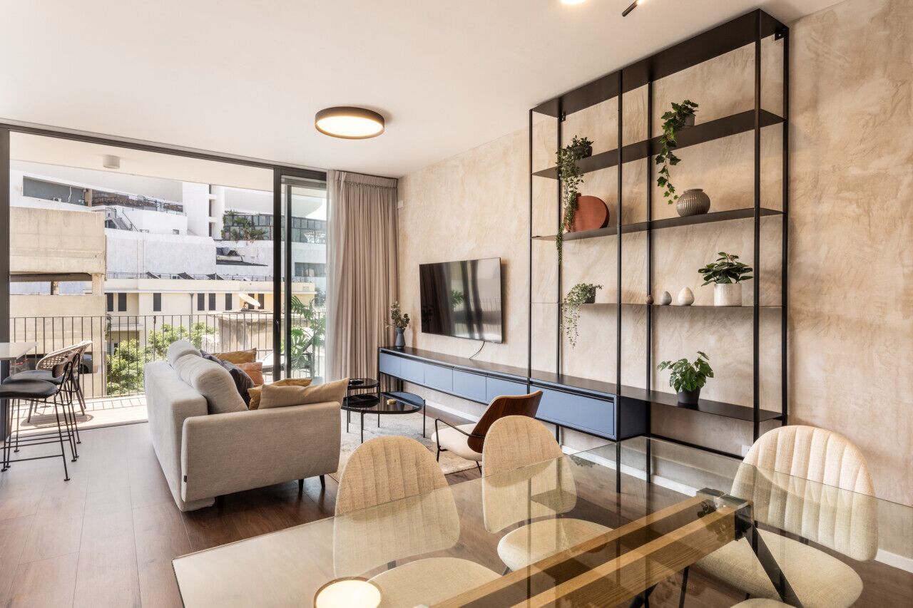 A modern living room with a sofa, chairs, a glass dining table, and a TV mounted on the wall. There is a balcony with a view of buildings outside.