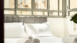 Una habitación de hotel con un ventanal grande, una cama bien hecha con ropa blanca y un cabecero gris.