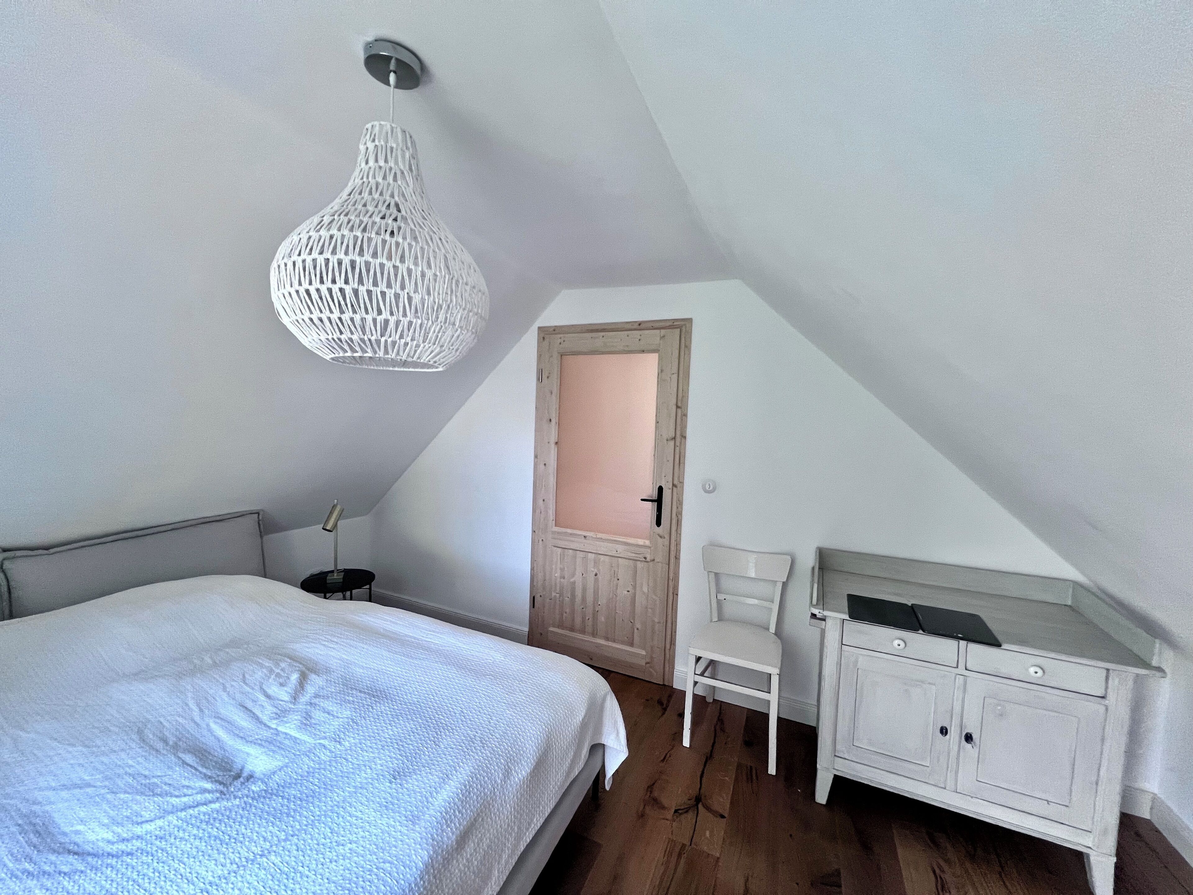 A bedroom with a bed, a white chair, a white dresser, and a white hanging light fixture.