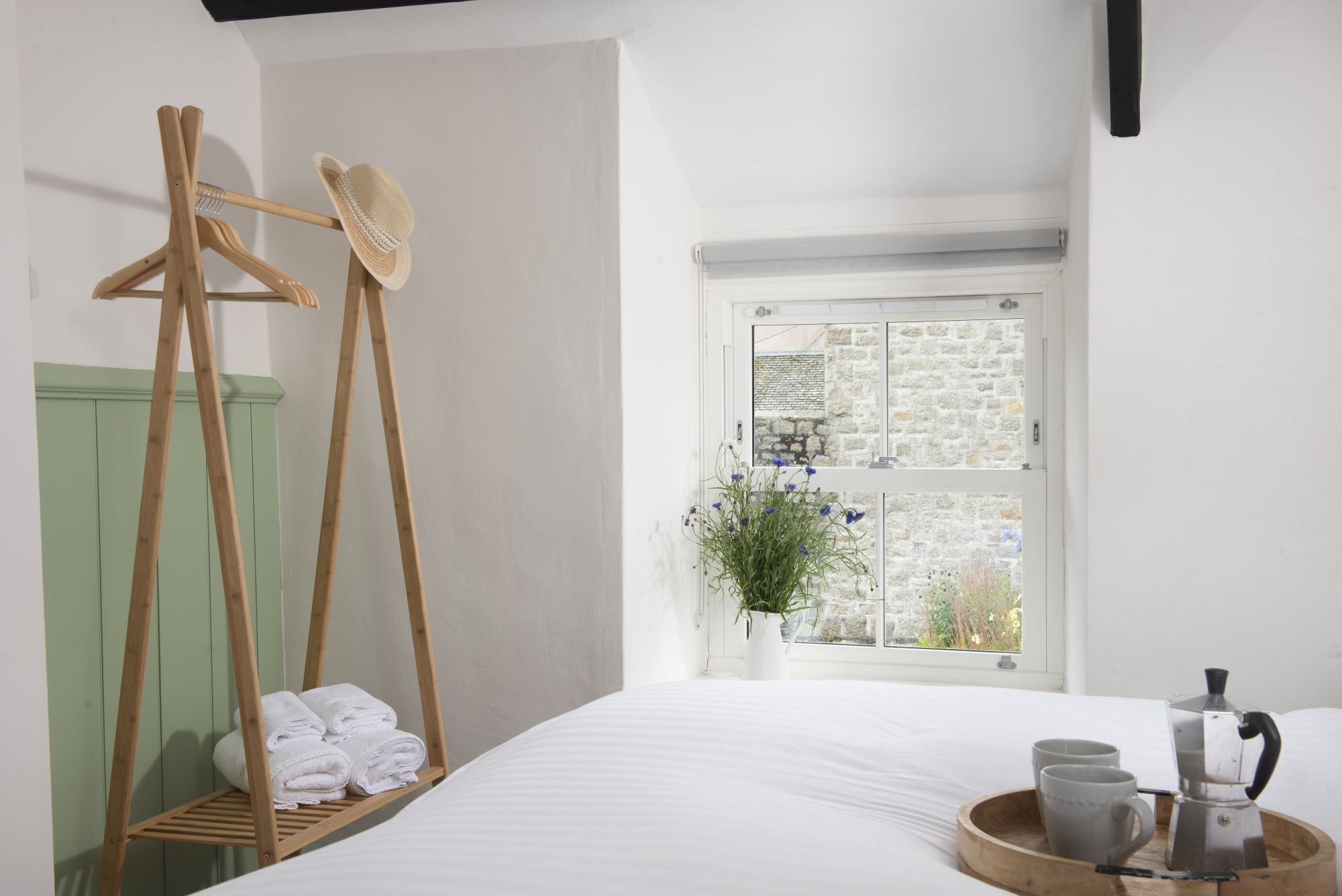 A bedroom with a bed, a wooden drying rack, a window with a view of a stone building, and a tray with a coffee maker.