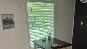 Een klein houten bureau met een stoel, een raam met zonwering, een fotolijstje en een plank met spullen.