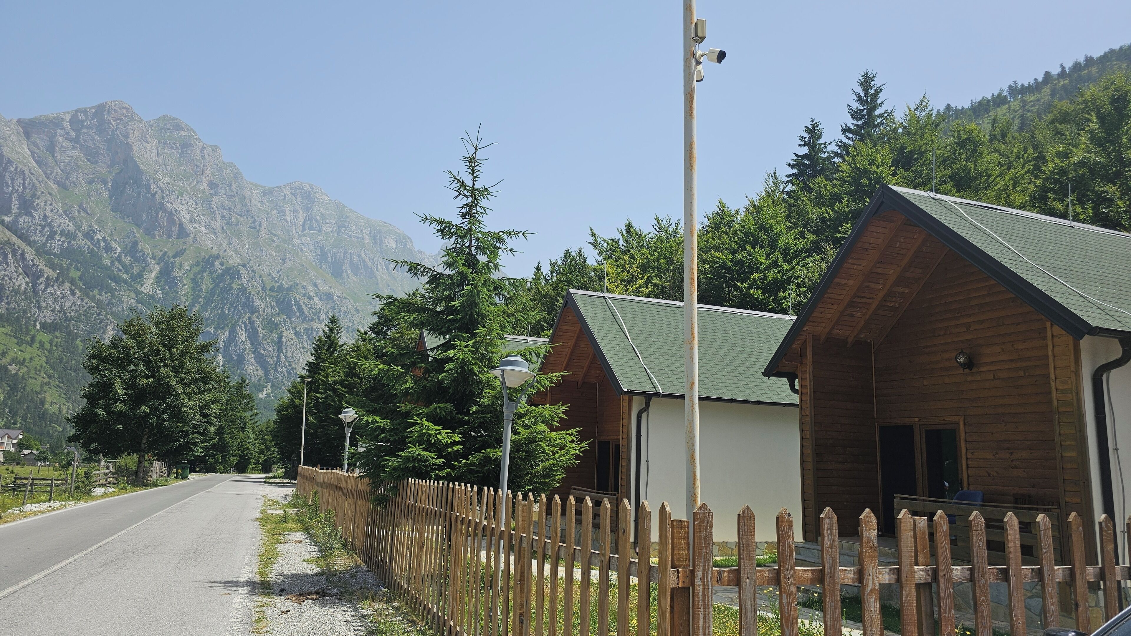 Une cabane en bois surmontée d’un toit vert, entourée d’une clôture en bois, avec un paysage montagneux en toile de fond.