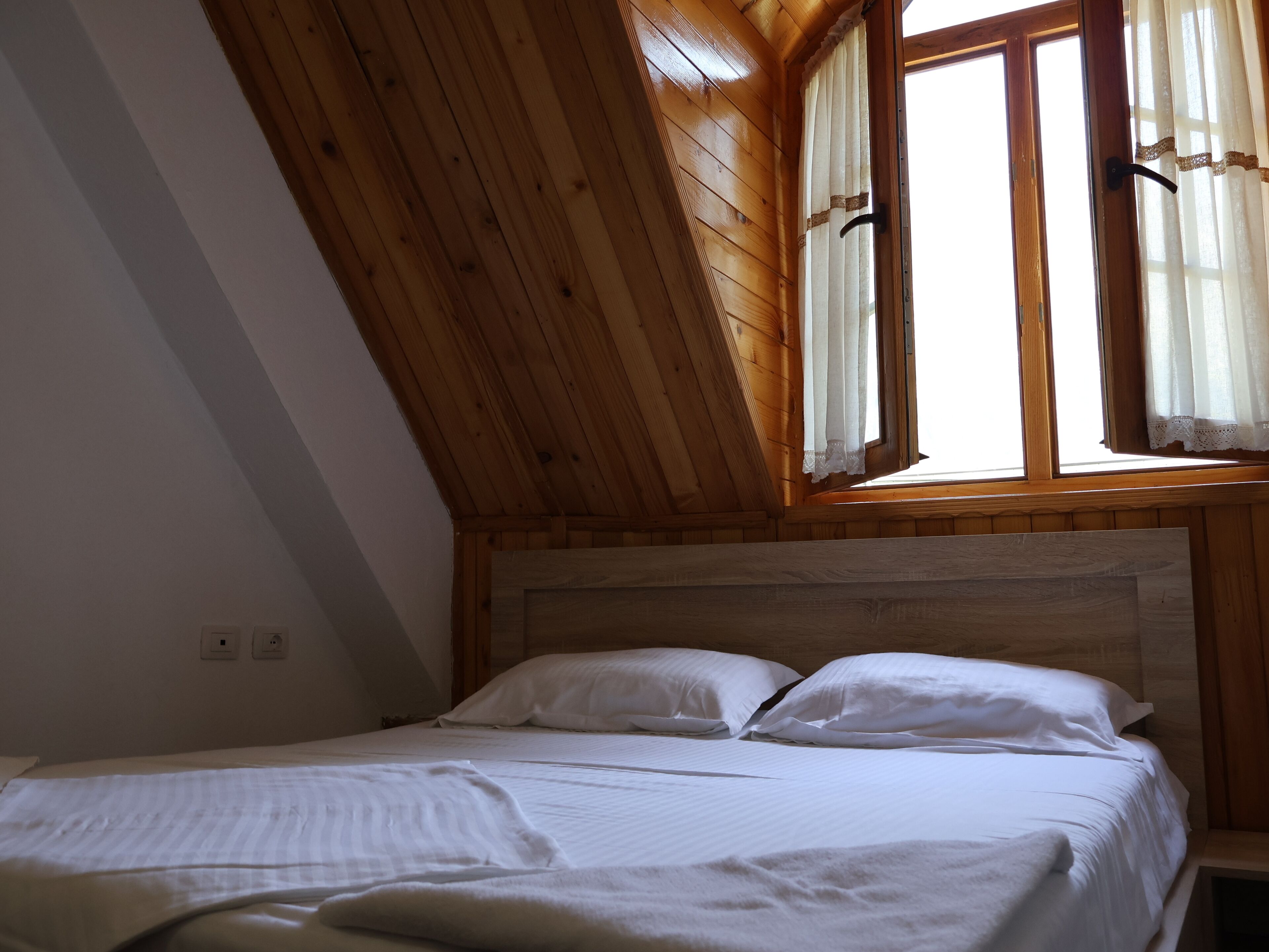 Une chambre avec un plafond en pente en bois, un lit avec une literie blanche et une fenêtre avec des rideaux.