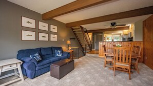 A cozy living room with a blue sofa, a wooden dining table with chairs, a staircase, and framed pictures on the wall. - New! Mountain View Chalet w/ Hot Tub & Fireplace (McHenry)