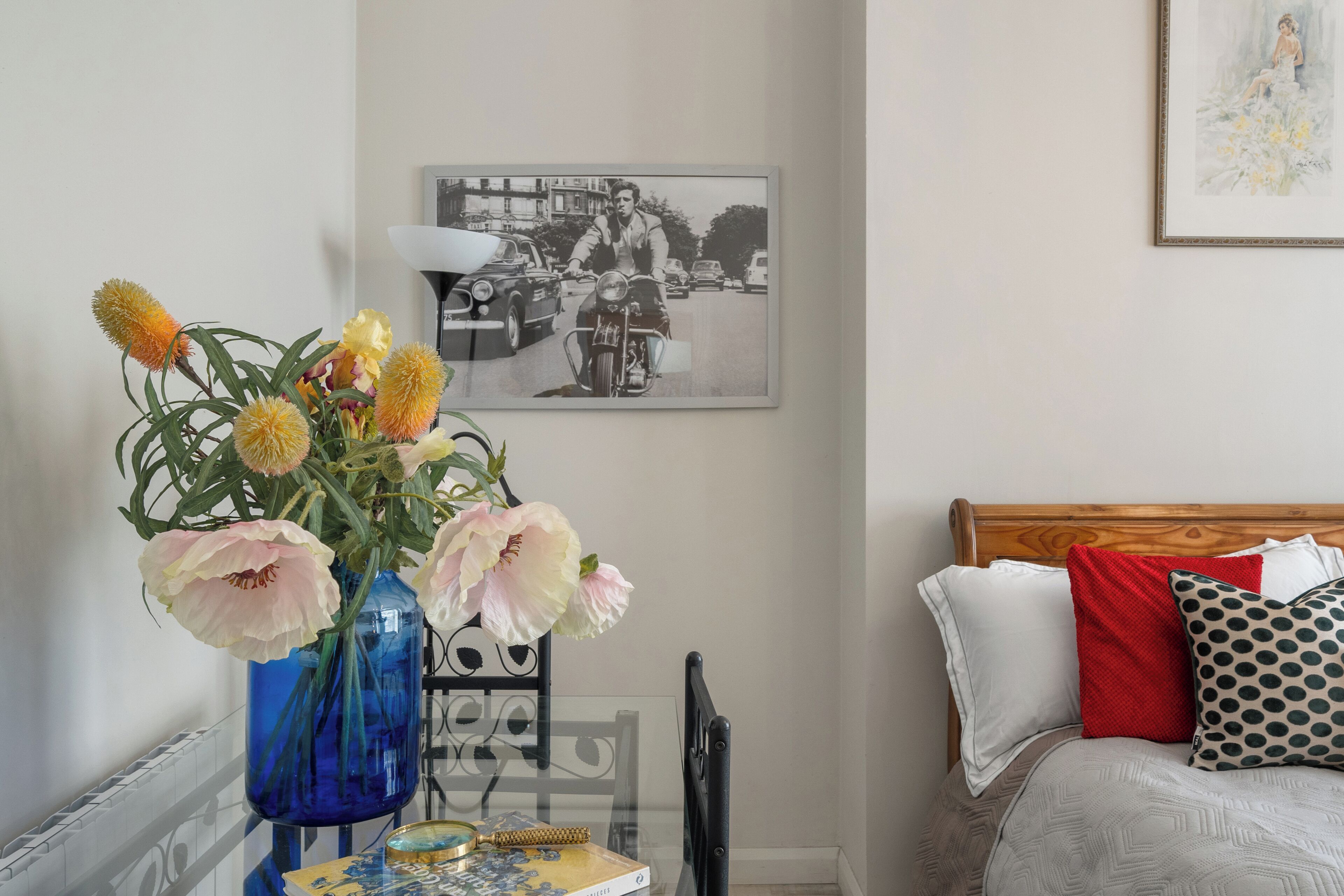 Un dormitorio con cama, almohadas, un florero con flores, un cuadro en la pared y una mesa con un libro y una lupa.